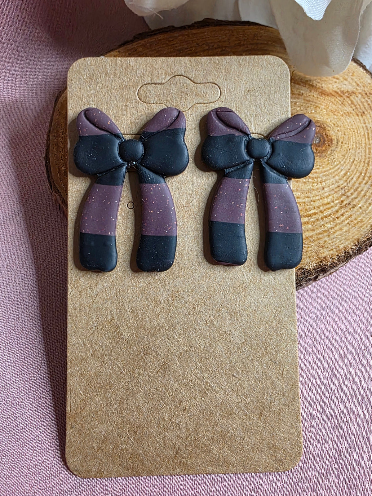 Handmade dark purple and black color block clay bow stud earrings with glitter shimmer on kraft earring card on wood slice