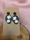 Handmade black clay dangle earrings with white ghost and skull appliqués on a kraft earring card